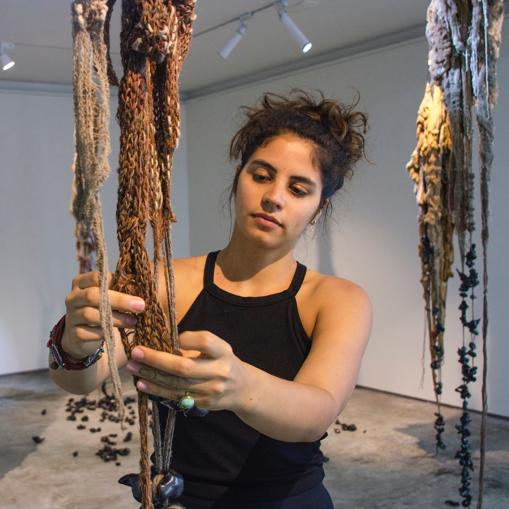 Romina wears tank top and manipulates fiber hanging from the ceiling